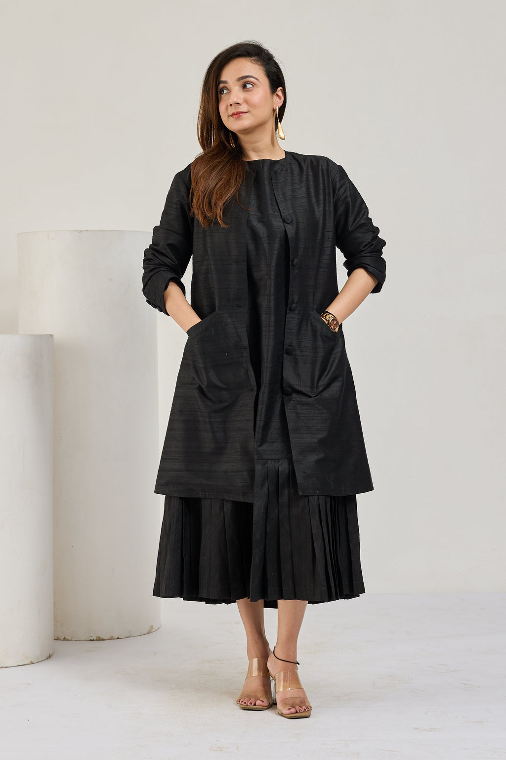 Model in black raw silk knee-length coat with front pockets over a pleated midi dress, looking to the left