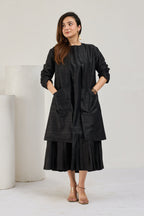 Model in black raw silk knee-length coat with front pockets over a pleated midi dress, looking to the left