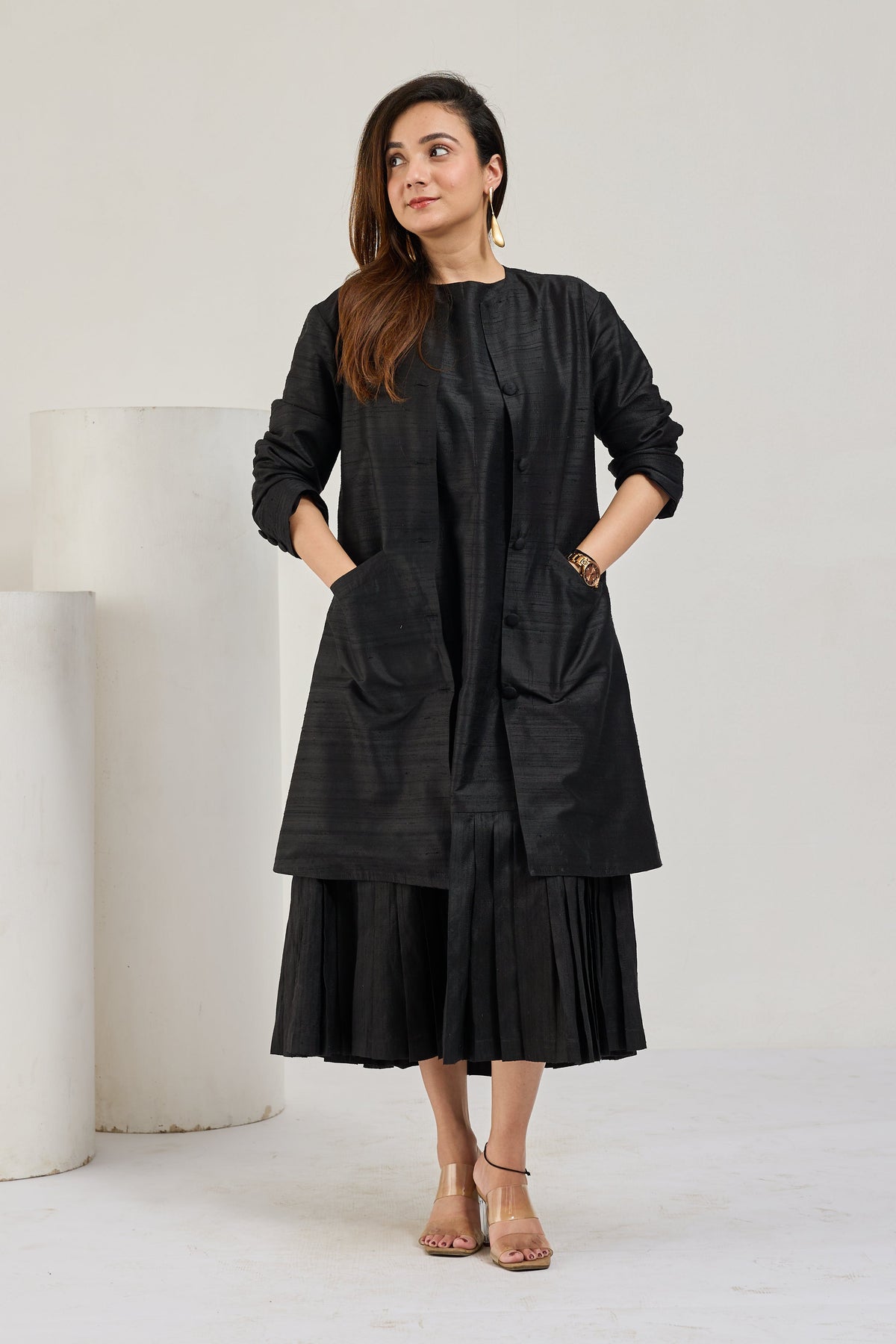 Model in black raw silk knee-length coat with front pockets over a pleated midi dress, looking to the left