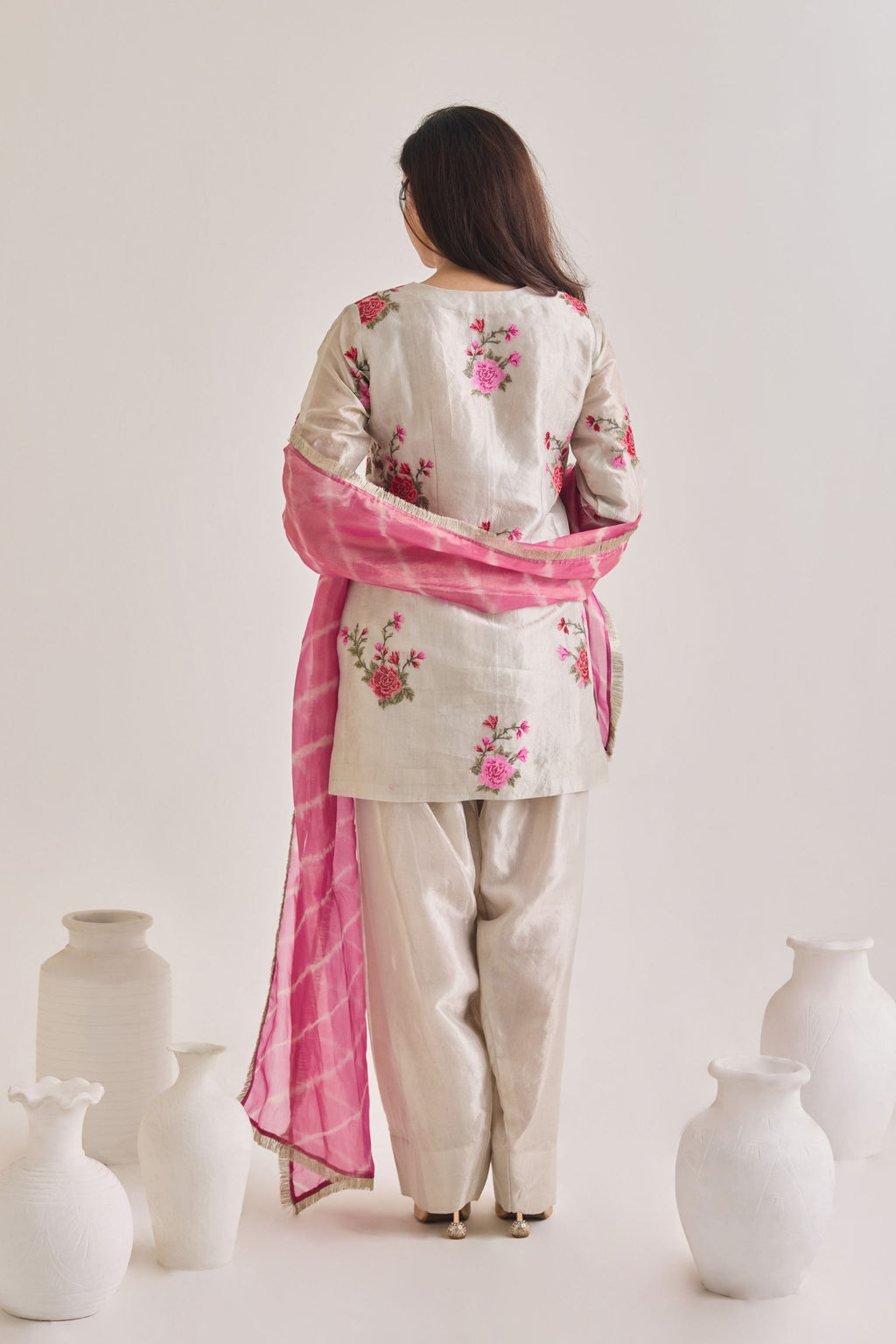 Back view of Silver and pink Farsi suit salwar set made out of Silver tissue with a machine embroidered rose motif kameez having 3/4th sleeves, a farsi salwar and Pink organza laheriya print dupatta paired with a Potli Bag.