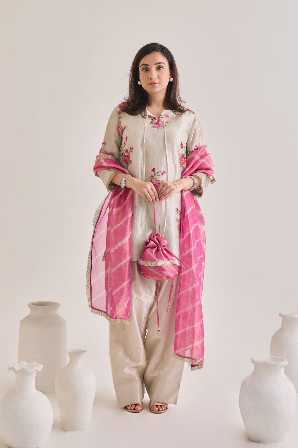 Front shot of Silver and pink Farsi suit salwar set made out of Silver tissue with a machine embroidered rose motif kameez having 3/4th sleeves and tie neck detail, a farsi salwar and Pink organza laheriya print dupatta paired with a Potli Bag.