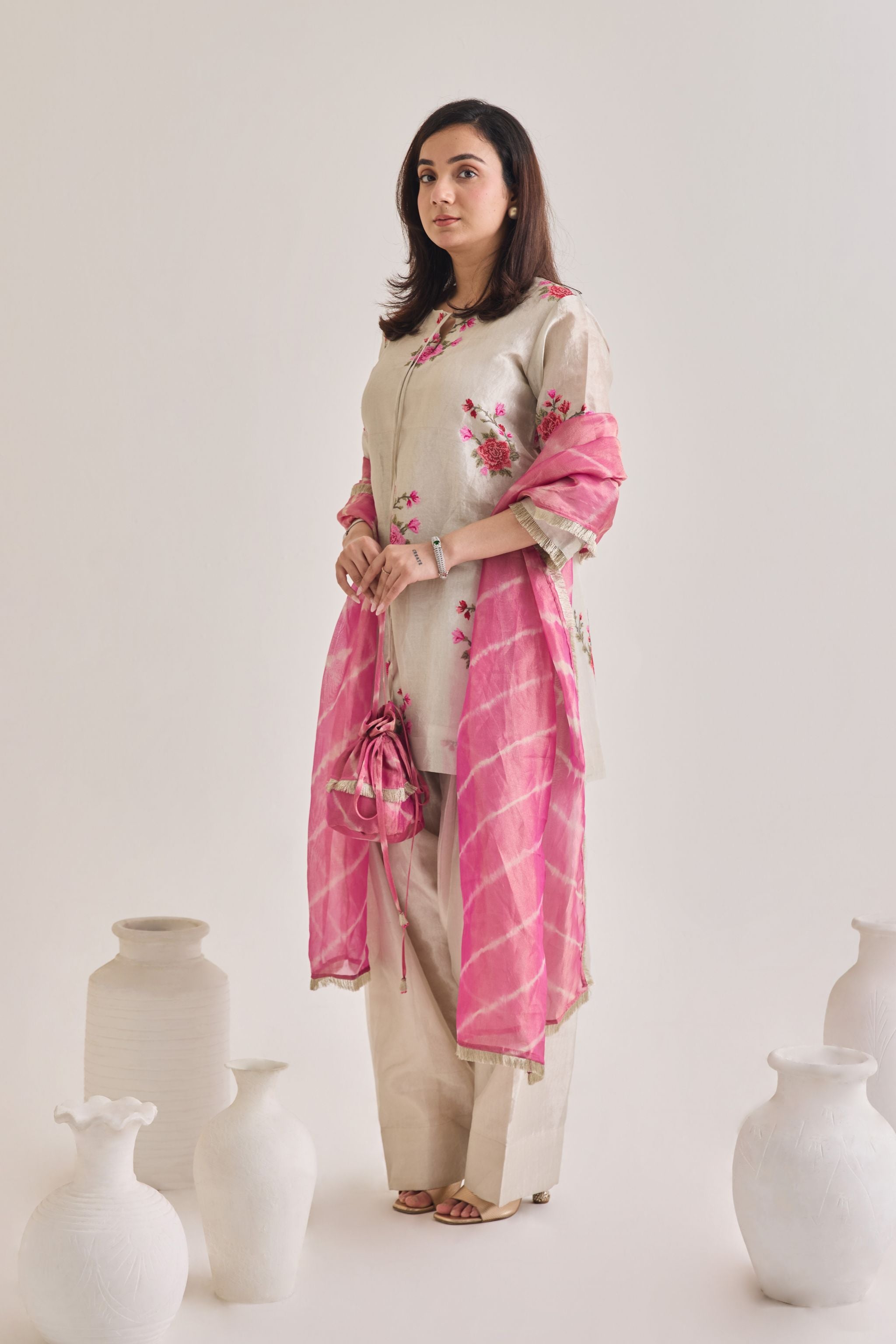 Side view of Silver and pink Farsi suit salwar set made out of Silver tissue with a machine embroidered rose motif kameez having 3/4th sleeves and tie neck detail, a farsi salwar and Pink organza laheriya print dupatta paired with a Potli Bag.